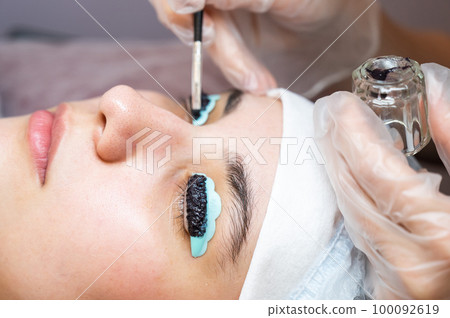 Close-up portrait of a woman on eyelash lamination procedure. The master applies tint to the eyelashes. Close-up portrait of a woman on eyelash lamination procedure. The master applies tint to the eyelashes. 100092619