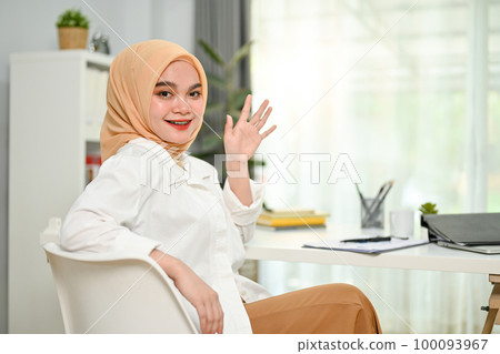 Cheerful Asian Muslim businesswoman smiling, waving hand, looking at camera, sitting at her desk. 100093967