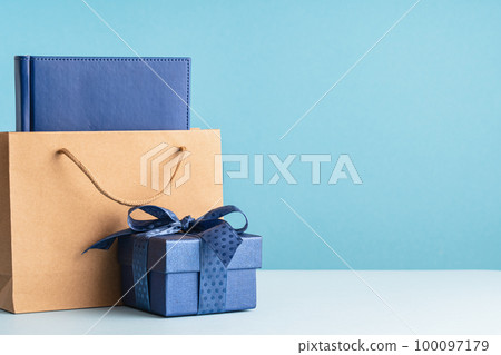 small Luxury gift box with a blue bow paper bag and agenda. Side view. Fathers day or Valentines day gift for him. Corporate gift concept or birthday party. Festive sale copy space small Luxury gift box with a blue bow paper bag and agenda. Side view. Fathers day or Valentines day gift for him. Corporate gift concept or birthday party. Festive sale copy space 100097179