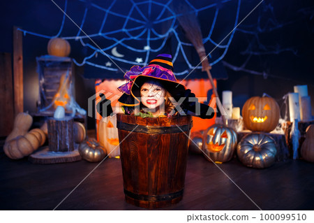 Little girl in witches costume scaring and making face while sitting in stupa on Halloween party 100099510