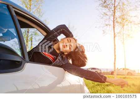 Happy woman waving her hand on an open windowed car against blue sky and sun. 100103575