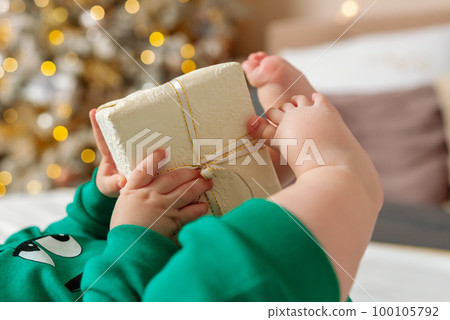 Baby gift. A little boy in a green suit holds a gift box with his feet and hands in bed. 100105792