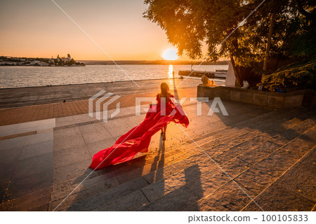 Sunrise red dress. A woman in a long red dress against the backdrop of sunrise, bright golden light of the sun's rays. The concept of femininity, harmony. 100105833