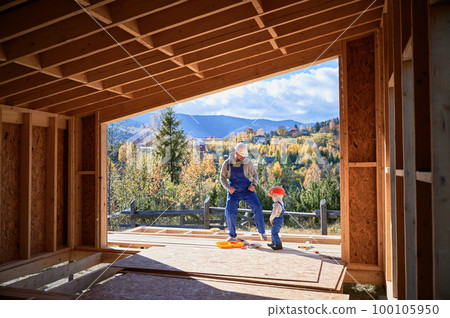 Father with toddler son building wooden frame house in the Scandinavian style barnhouse. Male builder having fun with kid on construction site on sunny day. Carpentry and family concept. 100105950