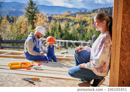 Father, mother and son building wooden frame house. Toddler boy helping his daddy, while woman looking for them on construction site. Guys wearing helmet and blue overalls. Carpentry, family concept. Father, mother and son building wooden frame house. Toddler boy helping his daddy, while woman looking for them on construction site. Guys wearing helmet and blue overalls. Carpentry, family concept. 100105951