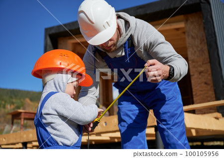 Father with toddler son building wooden frame house. Male builder and kid playing with tape measure on construction site, wearing helmet and blue overalls on sunny day. Carpentry and family concept. Father with toddler son building wooden frame house. Male builder and kid playing with tape measure on construction site, wearing helmet and blue overalls on sunny day. Carpentry and family concept. 100105956
