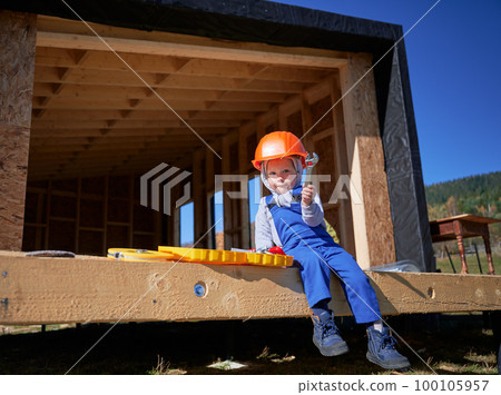 Boy toddler playing as builder on construction site. Child carpenter in orange helmet and blue overalls learning to build wooden frame house outdoor on sunny day. Carpentry and workshop concept. Boy toddler playing as builder on construction site. Child carpenter in orange helmet and blue overalls learning to build wooden frame house outdoor on sunny day. Carpentry and workshop concept. 100105957