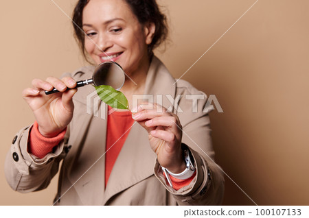 Selective focus on a green leaf in hands of blurred woman, attentively studying the structure of a plant through magnifying glass, on beige isolated background. Nature. Botany. Ecology. Free ad space Selective focus on a green leaf in hands of blurred woman, attentively studying the structure of a plant through magnifying glass, on beige isolated background. Nature. Botany. Ecology. Free ad space 100107133