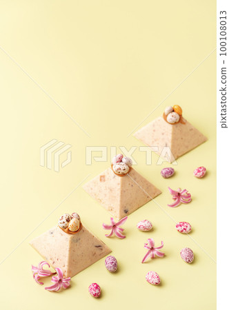 Traditional Easter cottage cheese dessert Paskha decorated with caramel and chocolate candy eggs and spring flowers on pastel yellow background. Modern minimalistic Easter background with copy space. Traditional Easter cottage cheese dessert Paskha decorated with caramel and chocolate candy eggs and spring flowers on pastel yellow background. Modern minimalistic Easter background with copy space. 100108013