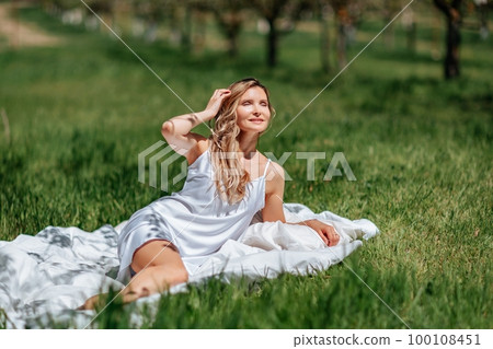 woman sleeps on a white bed in the fresh spring grass in the garden. Dressed in a blue nightgown. 100108451