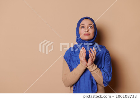 Peaceful Arab Muslim woman in blue hijab, looking up with gratitude, doing a day pray with folded hands, showing her believe in God isolated on beige backdrop. Islam. Religion. Middle-Eastern culture. Peaceful Arab Muslim woman in blue hijab, looking up with gratitude, doing a day pray with folded hands, showing her believe in God isolated on beige backdrop. Islam. Religion. Middle-Eastern culture. 100108738