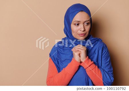 Middle-aged Muslim woman with head covered in blue hijab and hopeful look, keeping her hands folded on the chest level, praying Allah for peace, isolated beige background. Ad space. Close-up portrait. Middle-aged Muslim woman with head covered in blue hijab and hopeful look, keeping her hands folded on the chest level, praying Allah for peace, isolated beige background. Ad space. Close-up portrait. 100110792