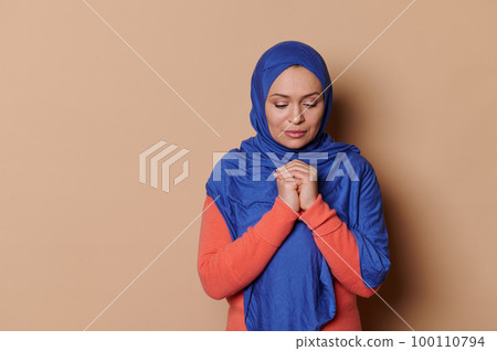 Middle-Eastern Muslim woman with head covered in blue hijab, holding her hands folded on chest level, praying for peace with hopefulness, isolated on beige background. People and Religion. Copy space 100110794