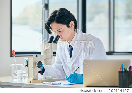 A young man in a white coat looking through the lens of a microscope and observing A young man in a white coat looking through the lens of a microscope and observing 100114197