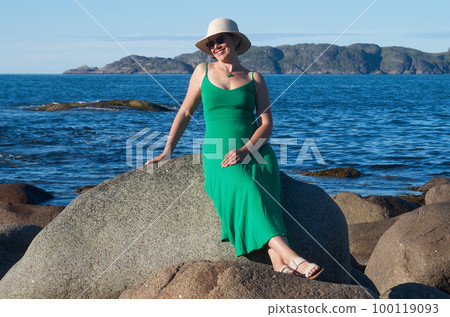 Woman tourist in a straw hat and a green dress on the seashore on a bright sunny day 100119093