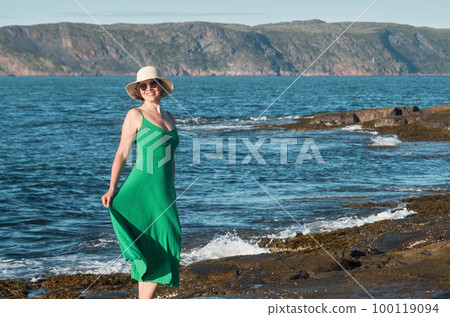 Woman tourist in a straw hat and a green dress on the seashore on a bright sunny day 100119094