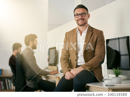 The expert in anything was once a beginner. Shot of a designer sitting on a desk with his colleagues blurred in the background. The expert in anything was once a beginner. Shot of a designer sitting on a desk with his colleagues blurred in the background. 100119389