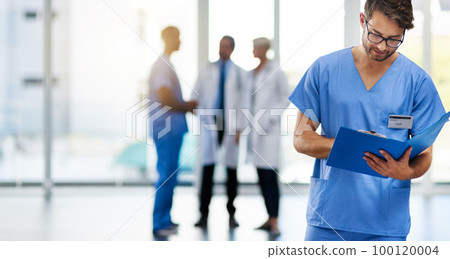 Young male doctor reading, holding and looking at patient health information at a hospital. Medical professional standing with doctors in the background. Healthcare worker working on a clinic file 100120004