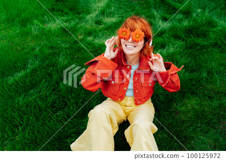 Happy smiling redhead woman with freckles in red orange gerbera Flower glasses sitting on green grass and listening to music in wireless headset. Positive Emotions. Fashion. Spring, summer mood 100125972