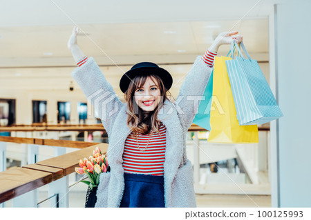 Happy, excited young fashion woman with raised hands holding paper shopping bags on mall background. Woman enjoying new spring season shopping. Consumerism, shopping, sale concept. Selective focus. Happy, excited young fashion woman with raised hands holding paper shopping bags on mall background. Woman enjoying new spring season shopping. Consumerism, shopping, sale concept. Selective focus. 100125993