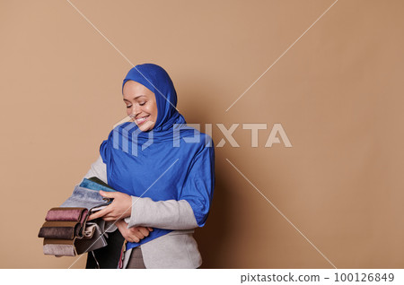 Multi-ethnic Muslim woman dressed in elegant gray suit and blue hijab, holding fabric samples of upholstery materials, isolated on beige color background. Interior design and home improvement conceptv 100126849