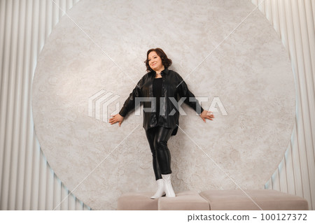 An adult woman in black leather clothes poses against a gray studio wall 100127372