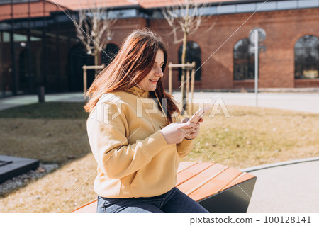 Beautiful woman in casual clothing using her smart phone while sitting on the bench on city street. Happy Young girl sitting on a bench after college classes. Urban lifestyle concept Beautiful woman in casual clothing using her smart phone while sitting on the bench on city street. Happy Young girl sitting on a bench after college classes. Urban lifestyle concept 100128141