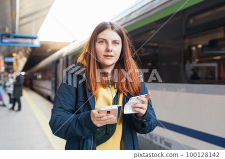 Young redhhead woman waiting on station platform with backpack on background electric train using smart phone. Railroad transport concept, Traveler 100128142