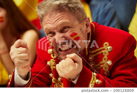 Close-up image of man, football, soccer fans emotionally watching match, worrying about favourite team. Fans wearing colorful scarves and flags Close-up image of man, football, soccer fans emotionally watching match, worrying about favourite team. Fans wearing colorful scarves and flags 100128809