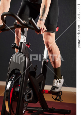 A young man on an exercise bike with a beard on a dark background.Training on an exercise bike in the gym. A young man on an exercise bike with a beard on a dark background.Training on an exercise bike in the gym. 100129821
