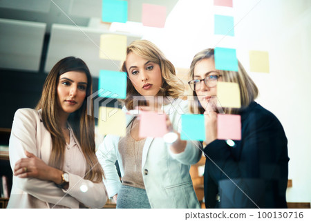 Talking team leader writing on sticky notes while planning, innovating or sharing business vision or ideas. Diverse group of motivated, ambitious or learning creative business women meeting together Talking team leader writing on sticky notes while planning, innovating or sharing business vision or ideas. Diverse group of motivated, ambitious or learning creative business women meeting together 100130716