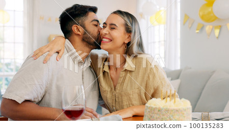 Interracial couple, gift and celebrate birthday being happy, kiss and smile in home at table with cake. Love, man and woman being content, romantic and present being cheerful celebration together. 100135283
