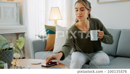 Carefree woman drinking coffee and feeling relaxed and refreshed while relaxing on the couch at home. Female taking deep breath and smelling the aroma of a hot beverage, feeling mindful and content 100135876