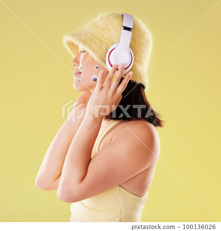 Makeup, music headphones and woman in studio isolated on a yellow background. Eye stickers, freedom technology and young female model listening, enjoying and streaming radio, podcast and audio song. 100136026