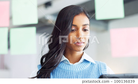 Focused young business woman thinking while brainstorming solutions for a project and marketing strategy in a creative startup agency. Designer planning ideas and information on a window in an office Focused young business woman thinking while brainstorming solutions for a project and marketing strategy in a creative startup agency. Designer planning ideas and information on a window in an office 100136839