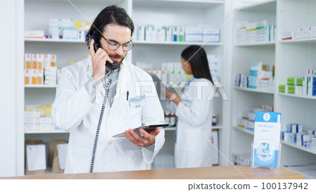 Pharmacist man answering the telephone and giving advice to customer on flu shot treatment options in pharmacy. Chemist assisting remote client by checking medicine stock on tablet 100137942