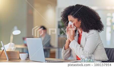 Sick businesswoman blowing nose with a tissue and suffering from flu virus or sinuses while working on a laptop in a modern office. Professional female with afro feeling ill while busy typing email 100137954