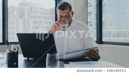 Businessman, office and drink water with documents, laptop and focus for market research, analysis or target. Advertising expert, paper or strategy with data analytics, kpi or goals for marketing job 100140311