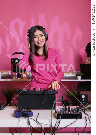 Artist standing at dj table mixing electronic sounds with techno using professional mixer console, having fun in studio over pink background. Asian musician playing stereo sounds with electronics Artist standing at dj table mixing electronic sounds with techno using professional mixer console, having fun in studio over pink background. Asian musician playing stereo sounds with electronics 100141238