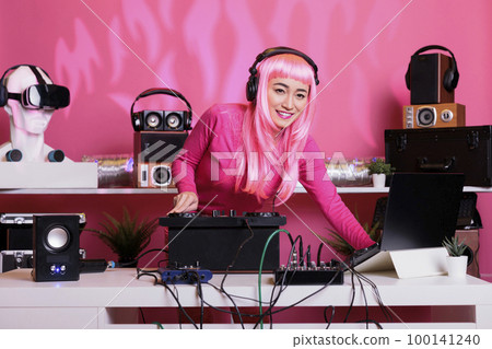 Asian musician standing at dj table wearing headset while playing techno music at professional mixer console in club at night time. Woman with pink hair having fun while enjoying performing song Asian musician standing at dj table wearing headset while playing techno music at professional mixer console in club at night time. Woman with pink hair having fun while enjoying performing song 100141240