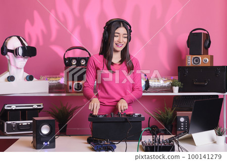 Asian artist performing electronic sound at professional mixer console in studio, enjoying mixing techno sound during concert. Cheerful performer playing stereo sounds with electronics and microphone 100141279