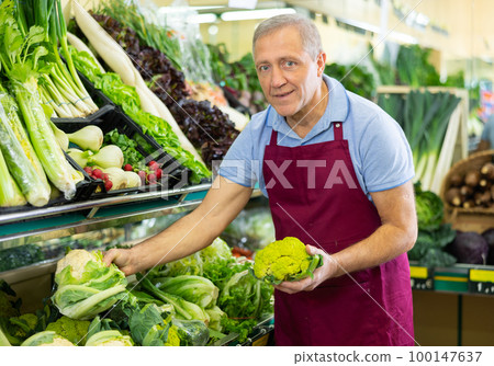Friendly mature salesman in apron arranging cauliflower in shop 100147637