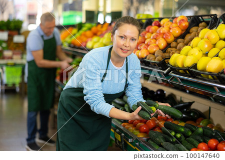 Middle-aged saleswoman putting cucumbers on food stall in greengrocery Middle-aged saleswoman putting cucumbers on food stall in greengrocery 100147640