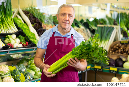 Aged salesman holding celery in front of food stall in greengrocery Aged salesman holding celery in front of food stall in greengrocery 100147711