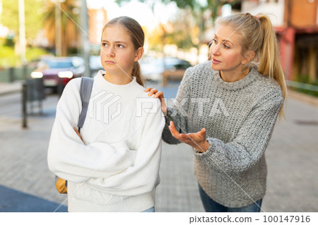 Frowning teenage girl listening to reprimanding from mother oncity street 100147916