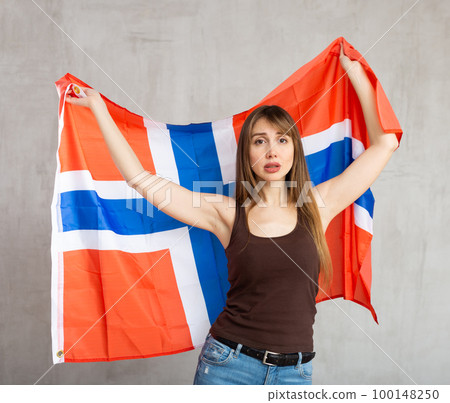 Upset patriotic young female student holding state flag of Norway against gray wall background indoors Upset patriotic young female student holding state flag of Norway against gray wall background indoors 100148250