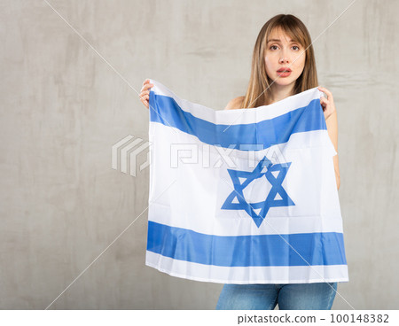 Sad young woman holding flag of Israel against unicoloured background 100148382
