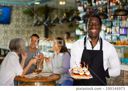 Positive African American waiter serving tapas for friends meeting for drinks and food in the pub 100148973