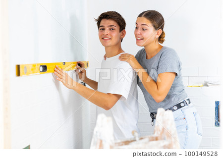 Positive couple checking laid tiles on wall with level meter in room under renovation 100149528