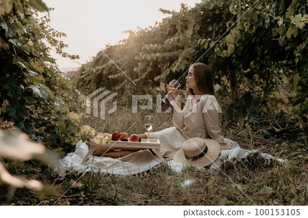 Woman picnic vineyard. Happy woman with a glass of wine at a picnic in the vineyard, wine tasting at sunset and open nature in the summer. Romantic dinner, fruit and wine. Woman picnic vineyard. Happy woman with a glass of wine at a picnic in the vineyard, wine tasting at sunset and open nature in the summer. Romantic dinner, fruit and wine. 100153105